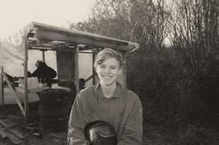 Joakim, 19, in blue coveralls holding a paintball mask at a paintball field.