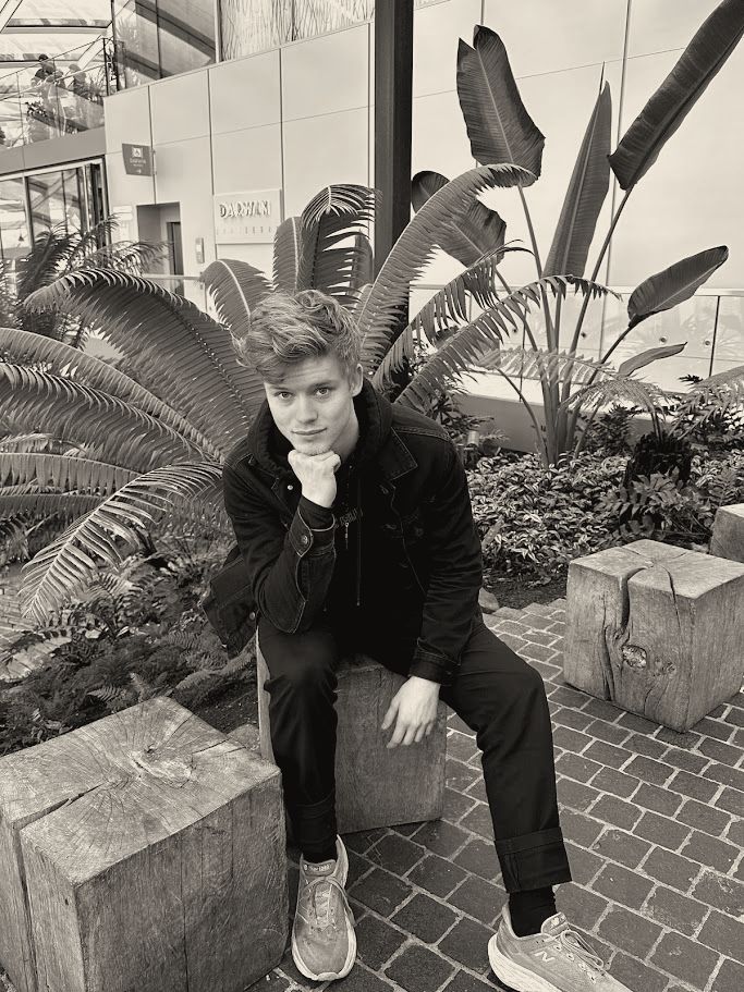 Joakim seated on a wooden block in an indoor garden, chin on hand, dressed in black.